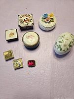 Eight decorative boxes on a pink surface showing variety including cat design, floral painted lids, porcelain and metal materials.