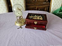 Photo showing both the ornate oriental jewelry box with detailed inlay artwork and the anniversary clock with glass dome on purple cloth surface.