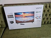 Front view of the boxed Vizio 24-inch D-Series Smart HDTV showing image of beach on screen and remote control.