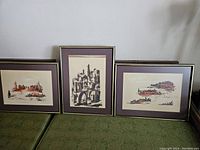 Three framed artworks featuring abstract cityscapes in reddish and black tones, matted and framed, arranged on a couch.