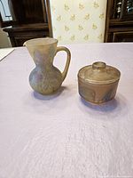 Pair of a ceramic pitcher and a covered bowl with frosted drip glaze and hand painted decorations on table.