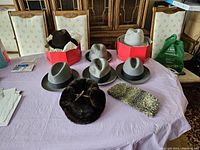Eight men's hats on lavender tablecloth: six felt fedoras in shades of gray and brown, one with feather band; two fur hats, one dark brown round, one elongated gray.