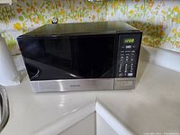Samsung microwave shown from front, placed on white counter, black door with stainless steel lower panel visible.