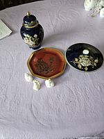 Overview of the collection showing the blue and gold lidded jar, black round plate, red octagonal tray, and three white figurines on a lavender table cloth.