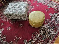 Two ottomans on a red patterned carpet: one square with floral fabric upholstery and one round yellow vinyl with embossed top