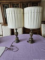 Pair of brass decorative table lamps with white pleated fabric shades on purple tablecloth in front of wooden cabinet.
