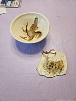 Top and angled views of a round pottery bowl and an irregular pottery wall plaque with hanging wire, both having cream base and brown brushstroke designs.