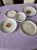5 decorative serving plates arranged on a lavender tablecloth, showing a variety of hand-painted fruit designs and a white lattice plate.