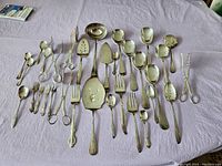 Full layout of 32 assorted silver plated serving utensils on a white cloth background showing variety of spoons, forks, and specialty pieces.