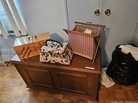 Photo showing three sewing-related storage items placed on a wooden chest: a wooden sewing kit with black handles on top, a floral fabric-covered sewing basket, and a geometric patterned knitting basket.