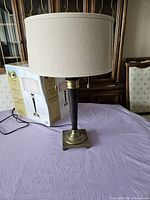 Front view of bronze finish table lamp with beige drum shade sitting on table with original packaging box behind it.