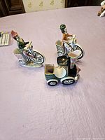 Trio of ceramic vintage kitsch figurines on a light purple cloth surface including two women on bicycles and a vintage car.