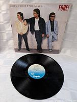 Color photo showing the vinyl record with the blue Chrysalis label resting on a white textured fabric background alongside the album sleeve featuring the three band members of Huey Lewis and The News.