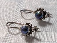 Close-up image of both sterling silver earrings laid flat on fabric, showing detailed silver Art Nouveau design surrounding central black pearls, lever-back hooks visible.