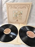 Two vinyl records on white paper sleeves displayed with the outer album cover behind them.