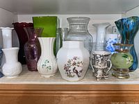 Shelf display of various vases in different colors and materials showing porcelain, hand cut and blown glass vases.