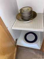 View of shelf with miscellaneous plates including a dark blue and gold-trimmed plate and a clear textured glass plate below the RS Germany bowl set.