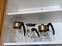 Three Mid-Century Modern glass coffee carafes displayed on a white shelf, showing the gold starburst patterns and black handles.