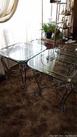 Two square end tables with bevelled glass tops and silver-tone metal bases with decorative scrollwork, placed indoors with carpet flooring and other furniture visible.