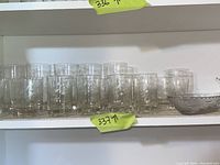 Three rows of clear drinking glasses of various sizes and patterns along with a glass serving dish on a white shelf.