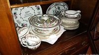 Photo showing stacked plates, teacups, covered vegetable bowl, gravy boat, and other pieces of Indian Tree china on wooden shelf.