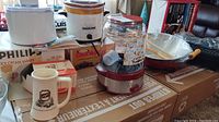 Wide view of lot on cardboard boxes showing popcorn maker, ice cream maker box, Rival Crock-Pot, Schlitz beer mug, and other items