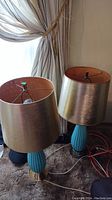 Two table lamps with ceramic bases and metallic gold cardboard shades placed on carpet in front of a curtain.