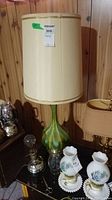 Front view of mid-century modern style table lamp with large cream fabric shade and green/gold swirled base on wood panel background.