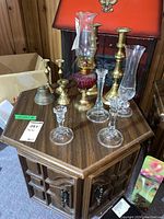 Wide view of all items on wooden table showing cranberry oil lamp, glass candle holders, brass candle holders, and brass bell.