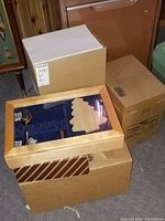 Three stacked cardboard boxes. Top box is wooden with transparent lid revealing liquor glasses inside. Other two boxes are plain cardboard likely containing paper supplies.