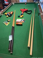 Full view of pool table with pool balls, snooker balls, pool cues, racks, cue chalk, instruction booklets, and brush arranged on felt surface