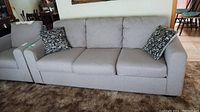 Full view of the gray upholstered sofa with three seat cushions, three back cushions, and two decorative patterned pillows on a carpeted floor.