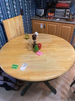 Round wood dining table with leaf installed, light natural wood tone top, dark green pedestal base, kitchen background.