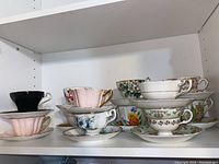 Eight assorted teacup and saucer sets arranged on a white shelf, showing various patterns and colors