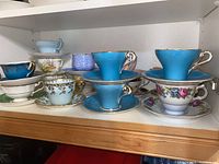 Multiple teacup and saucer sets on white shelf showing various colors and patterns
