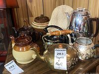 Photo showing seven assorted teapots of ceramic, metal and glass, plus quilted tea cozy with neutral color.
