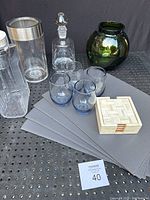 Overview photo showing 6 gray faux leather placemats, 4 blue vintage Libbey glasses, cream colored coaster box, clear glass decanter with stopper, green glass vase mark Poland, and a clear plexiglass wine chiller.