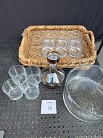 Entire lot showing 6 clear whiskey glasses placed in the sea grass basket, copper tone decanter in front, large round glass dish to the right.