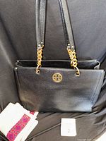 Front view of the black leather tote handbag showing gold-tone logo and chain handle detail. Dust bag partially visible.