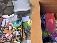 Overview of box contents showing various pens, pencils, a letter opener, and office tools placed in a shallow cardboard container and on desk.