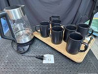 Photo shows the OXO electric kettle on a black base with power cord, seven matte black mugs arranged on a rectangular rustic wooden tray.
