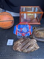 Photo showing two vintage worn baseball gloves, an NCAA signed basketball, a Wilson football in a wooden display case, and a mini basketball backboard with net placed on a black perforated surface.