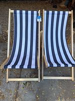Pair of wooden framed lounge chairs with blue and white vertical stripe canvas seats, positioned side-by-side on outdoor surface with dried leaves around.