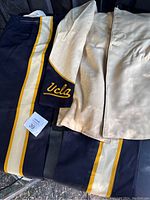 Photo showing one ivory jacket and two pairs of dark blue marching band pants with gold and white side stripes.