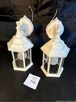 Two white exterior wall lantern lights with glass panels and bulbs, with wall mount backing and wires exposed.