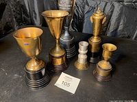 Front view of six antique trophies of various sizes and materials, including gold-tone, brass, and silver-tone with wooden or metallic bases.