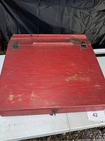 Red wooden writing desk box showing top surface with handle and metal latches. Visible surface scratches and worn paint.