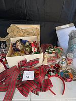 Box with assorted Christmas ornaments, large plaid ribbon bow, angel figurine, reindeer headband, and gift wrap rolls in background.