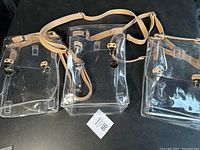 Three clear PVC messenger bags with beige adjustable straps displayed flat on a black surface, showing front view and closure details.