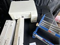 Wide shot showing Epson ET-2720 printer, Uline tape dispenser, toner boxes, and cords with light plugs on black surface.